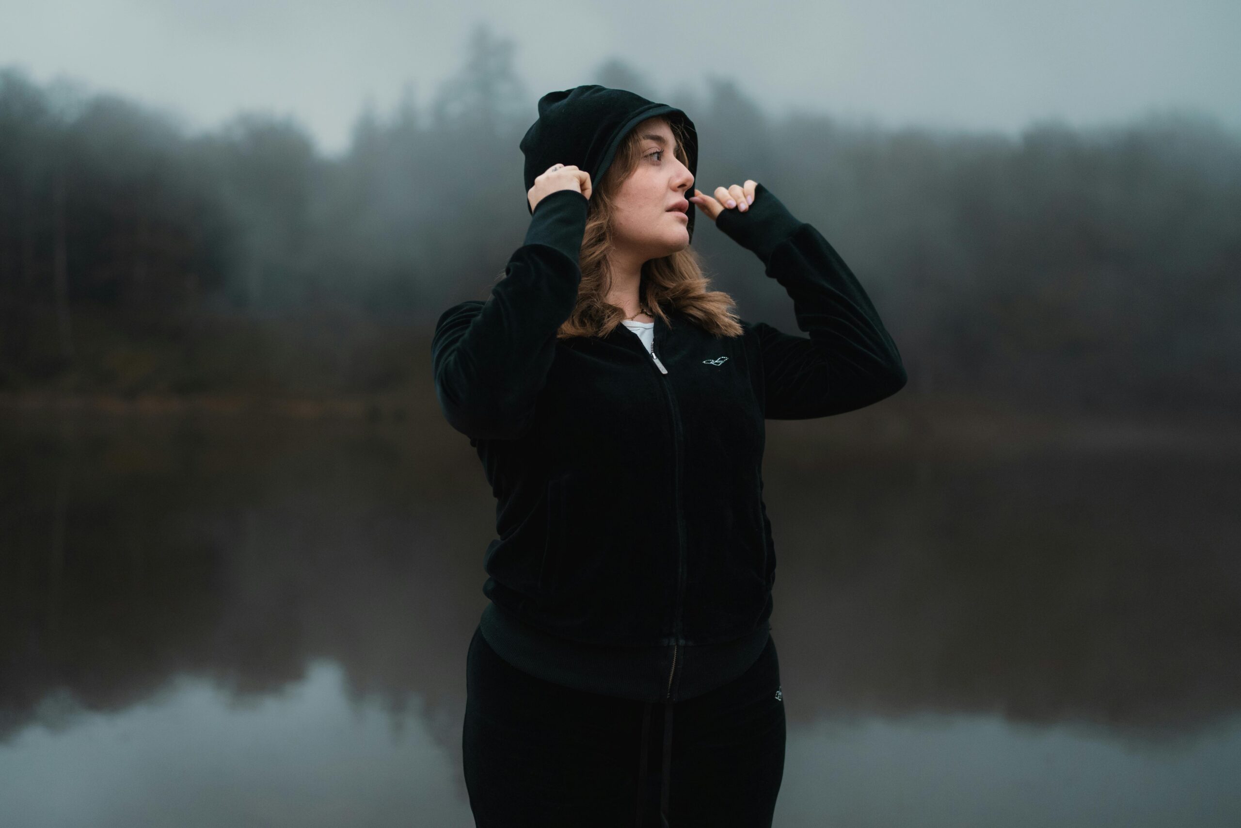 A young woman wearing a black hoodie stands by a misty lake, creating a serene and mysterious atmosphere.